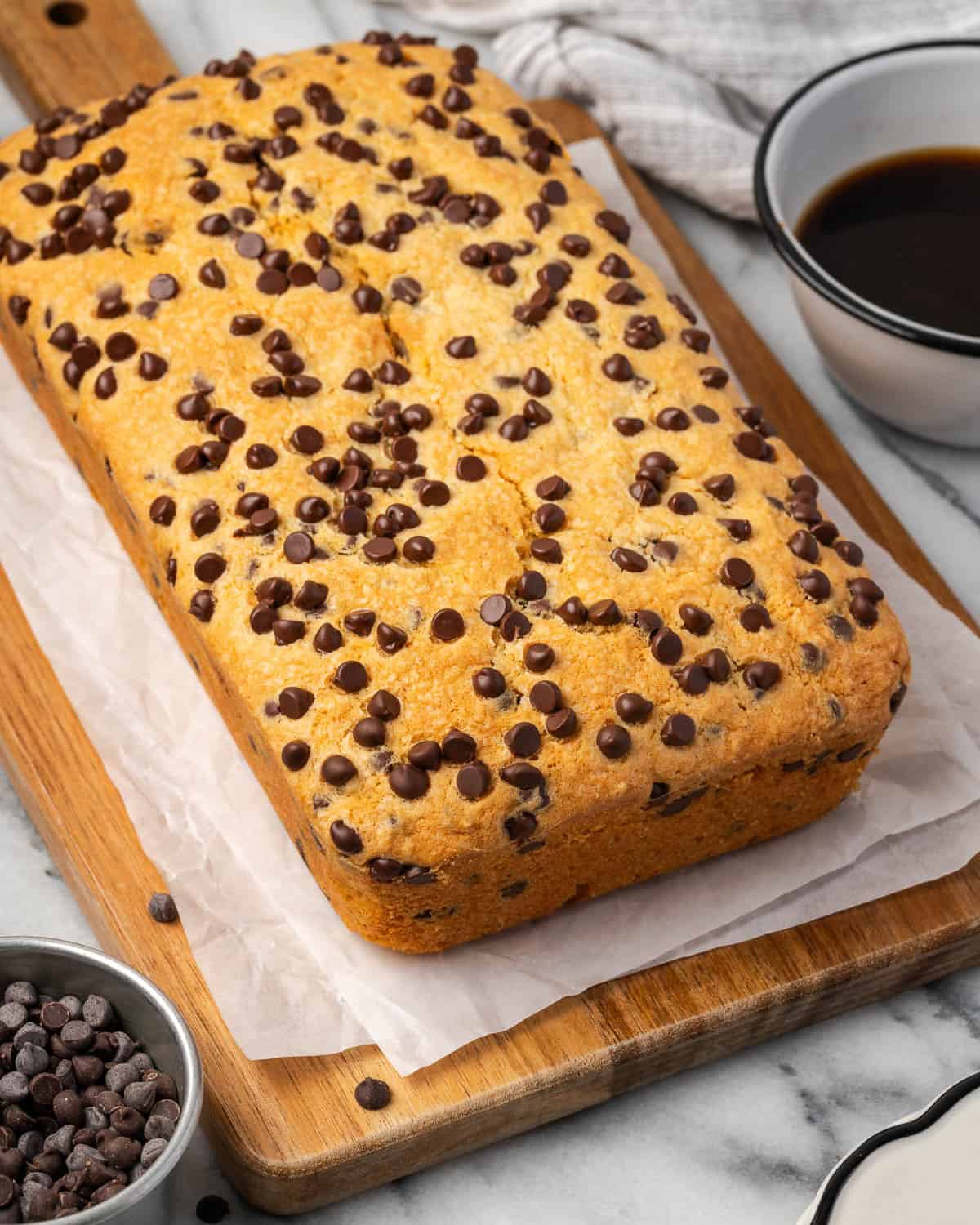 A freshly baked chocolate chip pound cake sitting on a cutting board next to a cup of coffee.