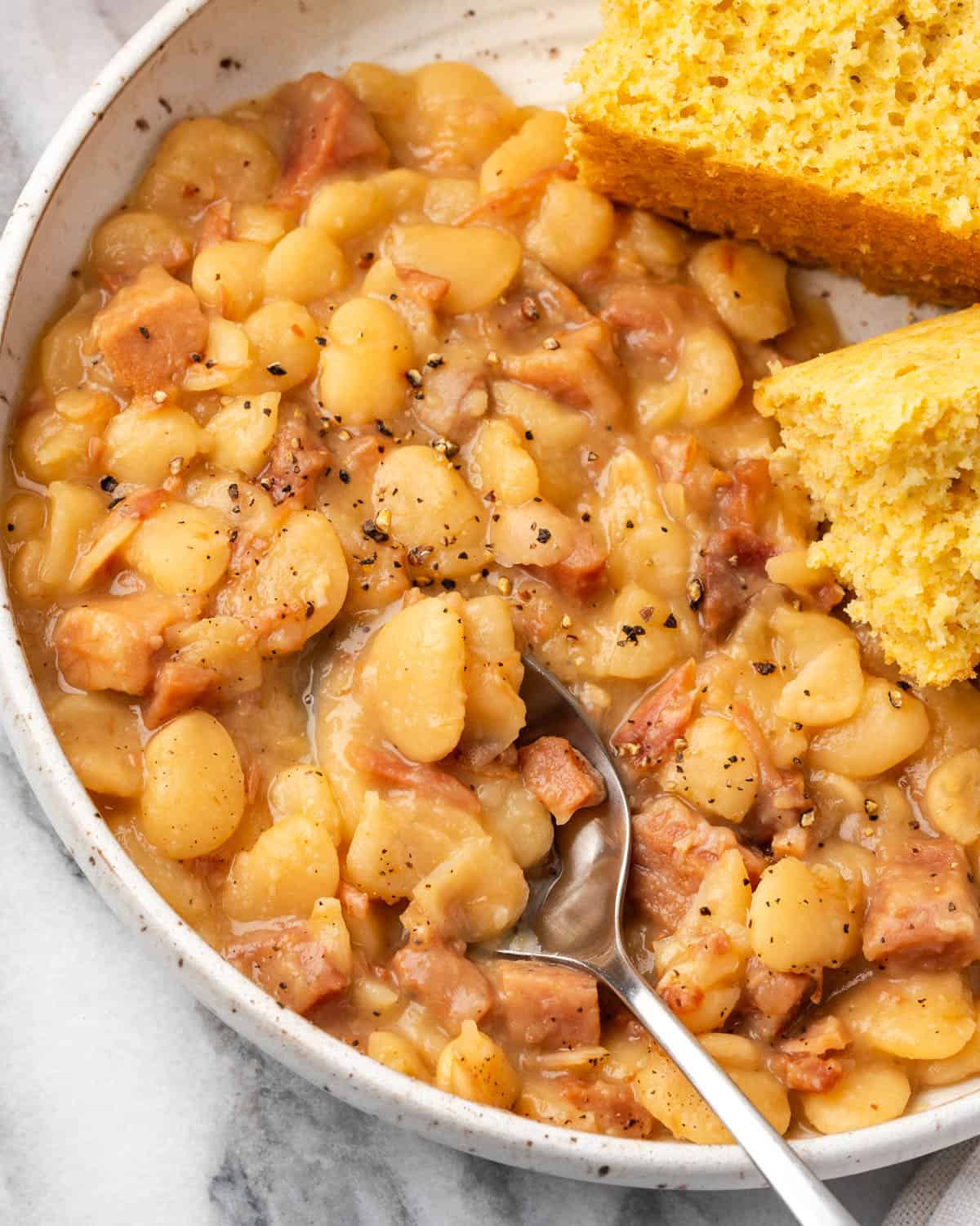 A spoon scooping up a bite of lima beans and ham from a serving plate. 