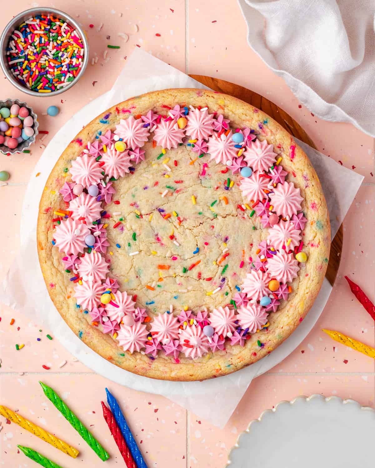 A sugar cookie cake sitting on a pink table next to bowls of sprinkles and assorted birthday candles.