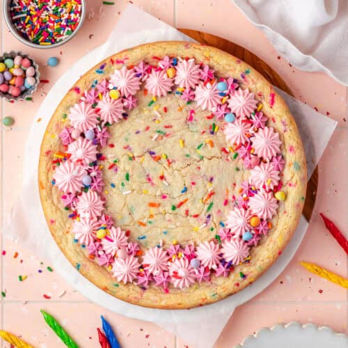 A sugar cookie cake decorated with pink frosting and sprinkles sitting on a pink tile table.