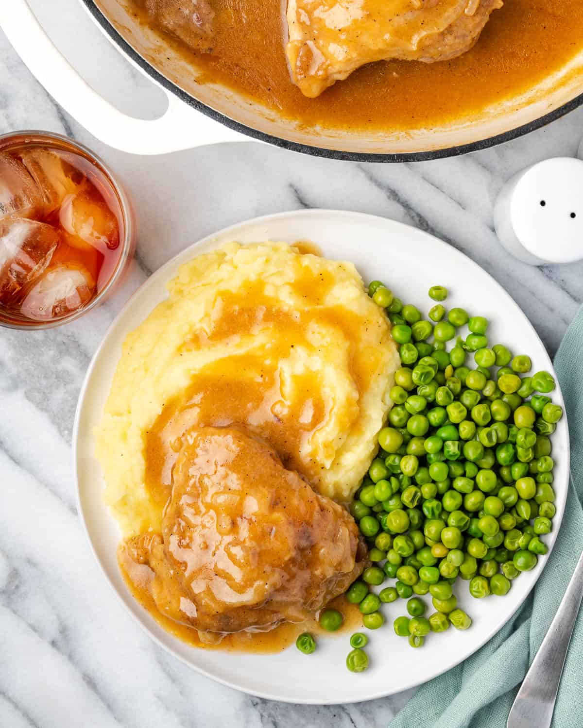 Smothered chicken thighs, mashed potatoes and peas on a dinner plate sitting next to a glass of iced tea.