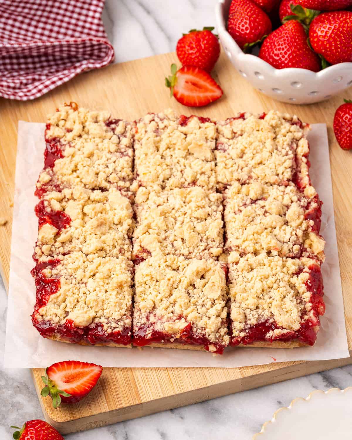 A batch of strawberry crumble bars cut into squares sitting on a cutting board next to a bowl of strawberries.