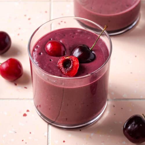 A cherry smoothie served in a small glass sitting on a tile counter.