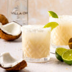 Two coconut lime margaritas sitting on a counter next to a bottle of tequila.