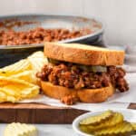 A sloppy joe sandwich sitting on a wood board next to some potato chips.
