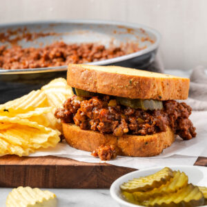 A sloppy joe sandwich sitting on a wood board next to some potato chips.