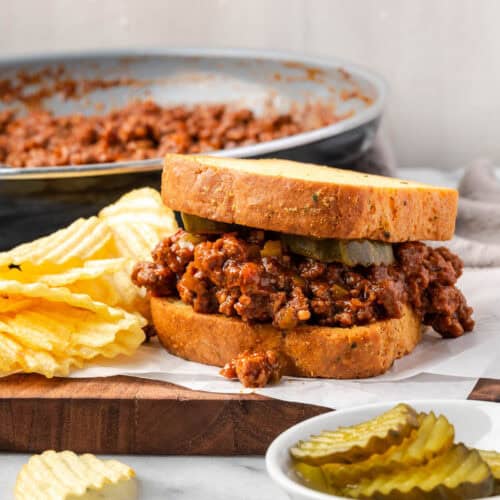A sloppy joe sandwich sitting on a wood board next to some potato chips.