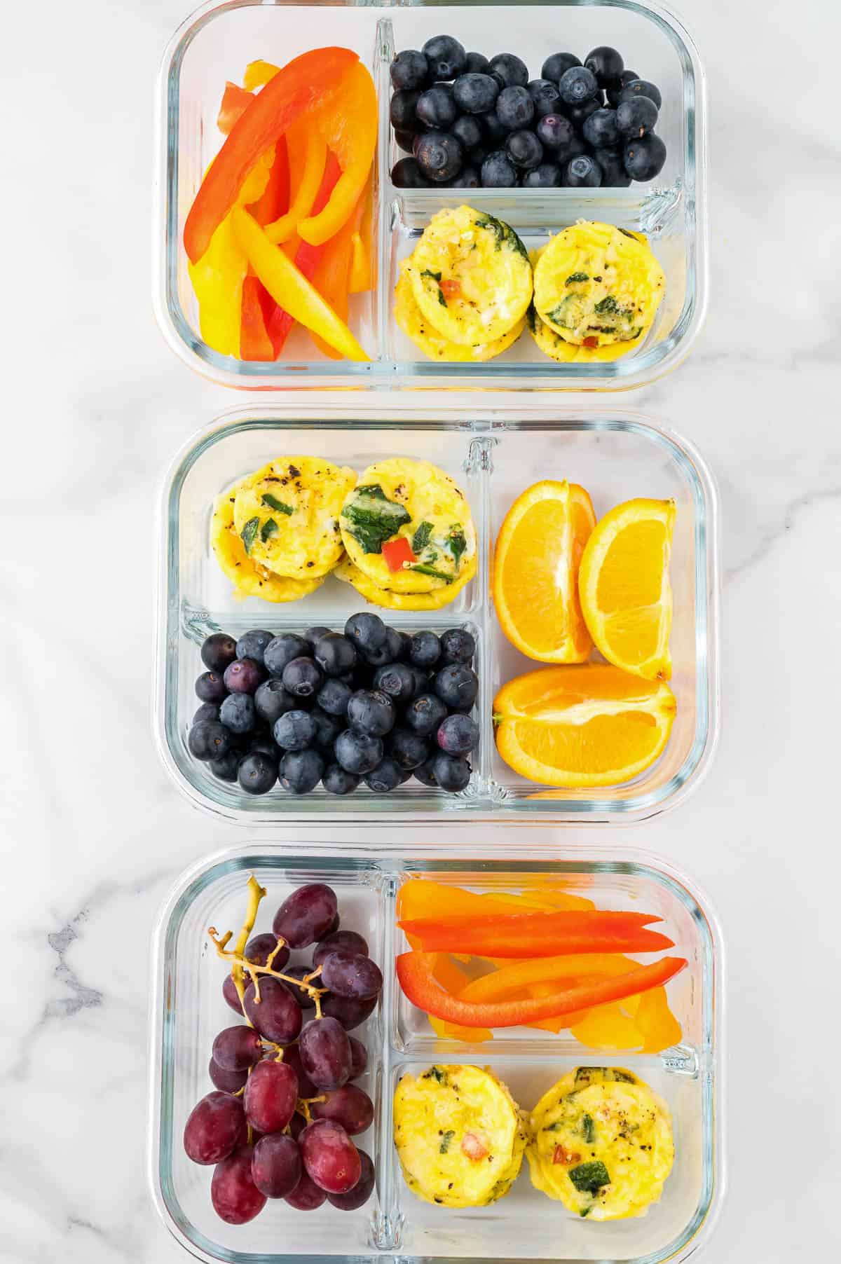 Three glass meal prep containers with egg bites, fruit and vegetables sitting on a marble counter.