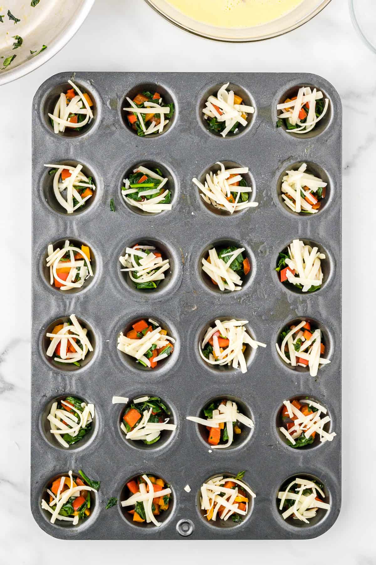 Cooked vegetables and shredded cheese portioned into a mini muffin tin.