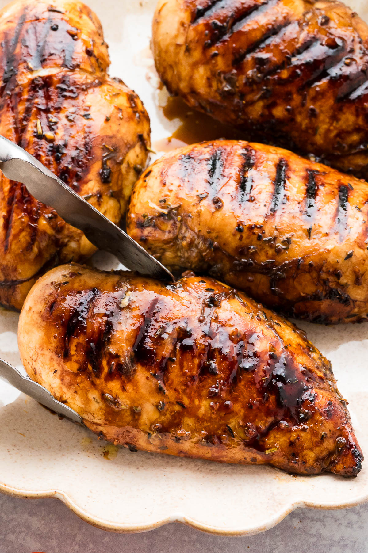 Tongs picking up a grilled piece of balsamic chicken from a serving plate.